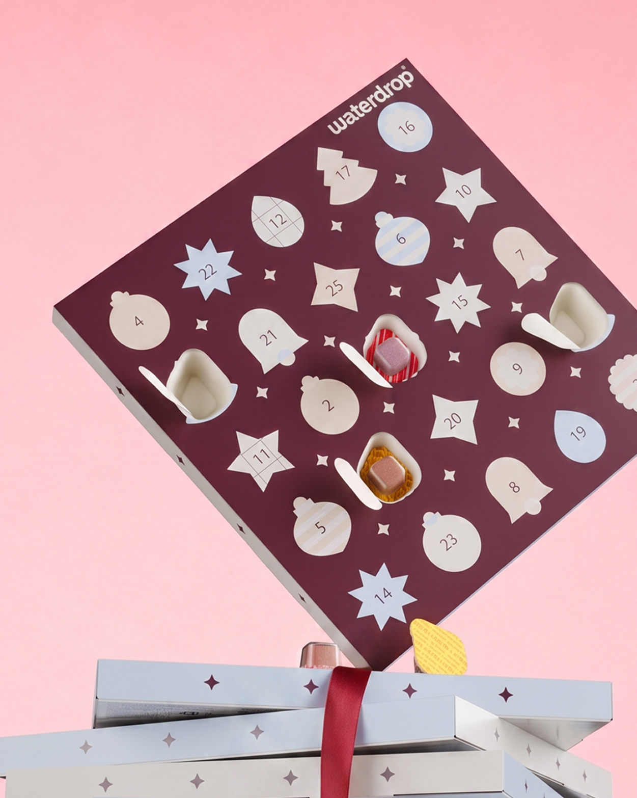 Purple Advent calendar with numbered star-shaped doors on a pink background, placed above two gift boxes.