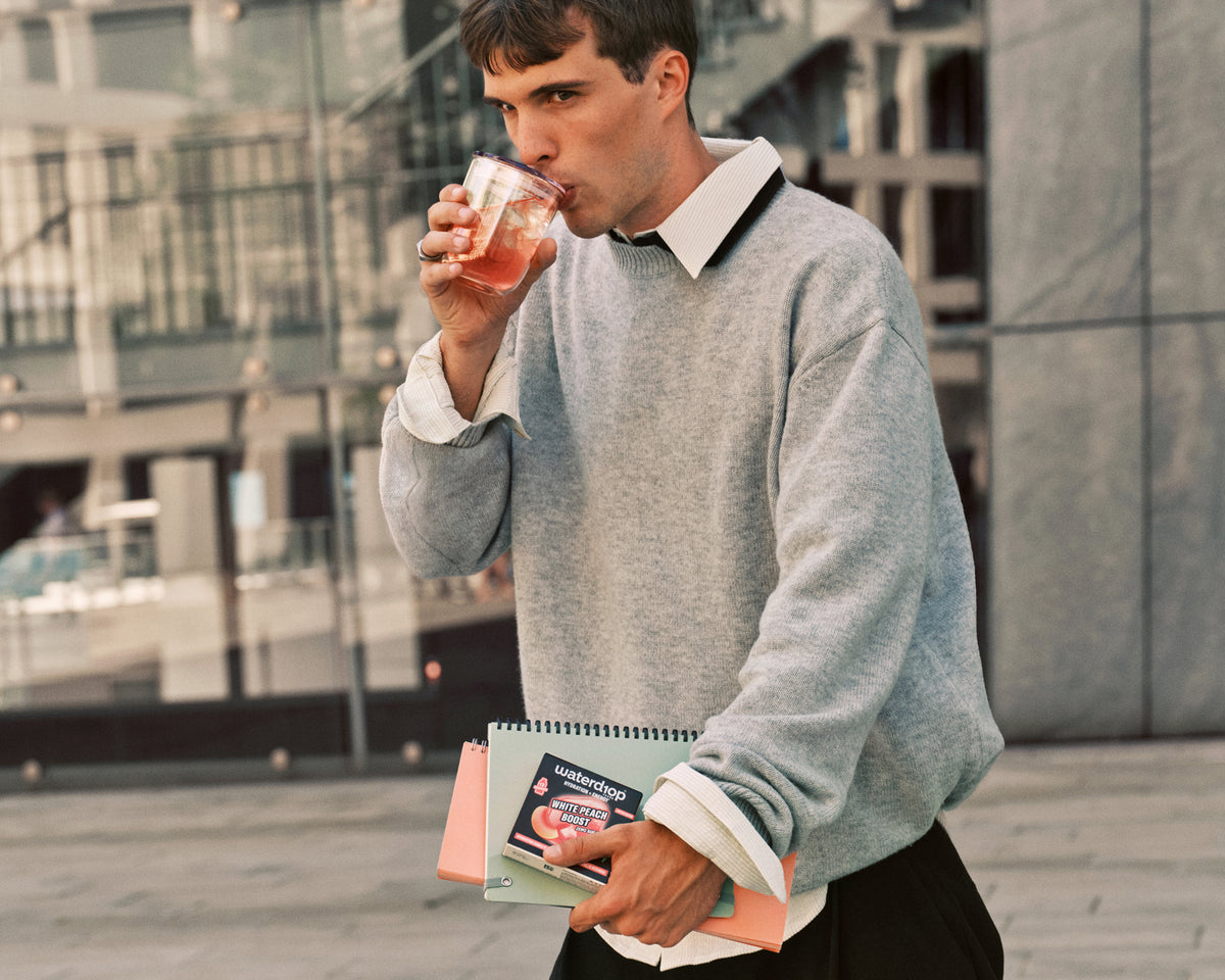 Man in a gray sweater drinks from a glass while holding a notebook outdoors in an urban setting.